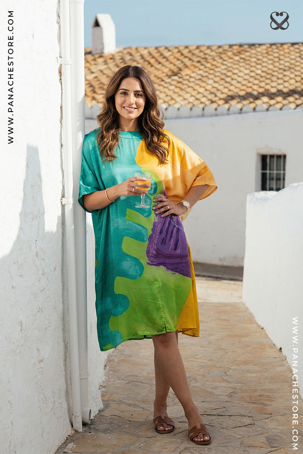 Woman in a colorful dress standing on a sunlit street with white walls and a blue sky.