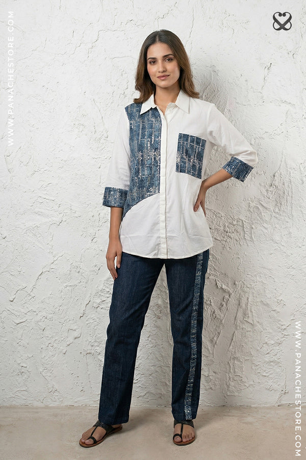 Woman wearing a white and blue patterned shirt with dark pants against a textured white wall.